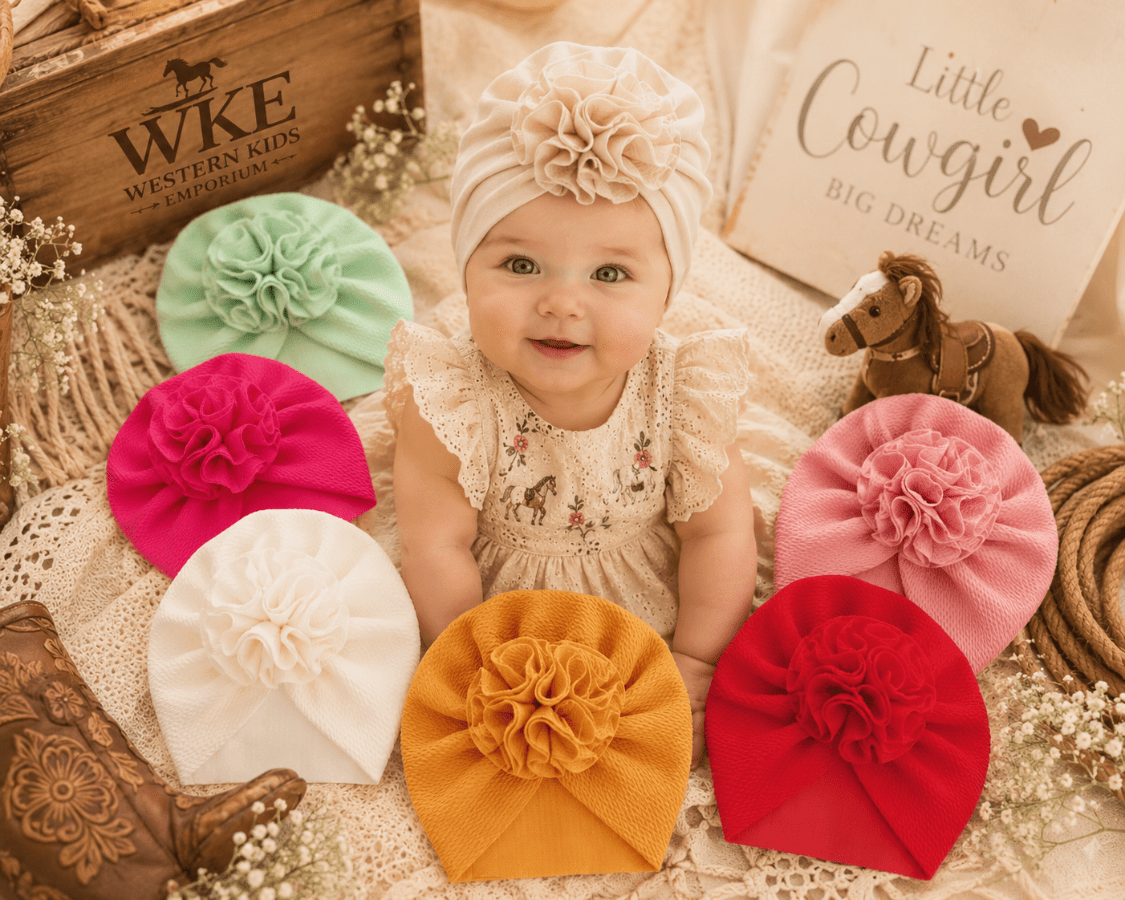 A baby wearing a cream ruffled bonnet with a large fabric flower sits on a textured blanket surrounded by six matching flower bonnets in mint green, magenta, white, mustard yellow, pink, and red. The scene includes western-themed props, including a wooden boot, a horse toy, and a sign reading 'Little Cowboy Big Dreams'.