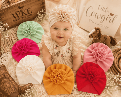A baby wearing a cream ruffled bonnet with a large fabric flower sits on a textured blanket surrounded by six matching flower bonnets in mint green, magenta, white, mustard yellow, pink, and red. The scene includes western-themed props, including a wooden boot, a horse toy, and a sign reading 'Little Cowboy Big Dreams'.
