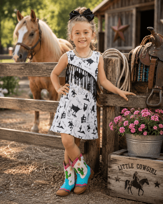 A young girl stands in a rustic barn setting wearing a sleeveless white dress with a black western-themed print and colorful cowboy boots. A horse is visible behind a wooden fence, and a bucket of pink flowers sits beside her.