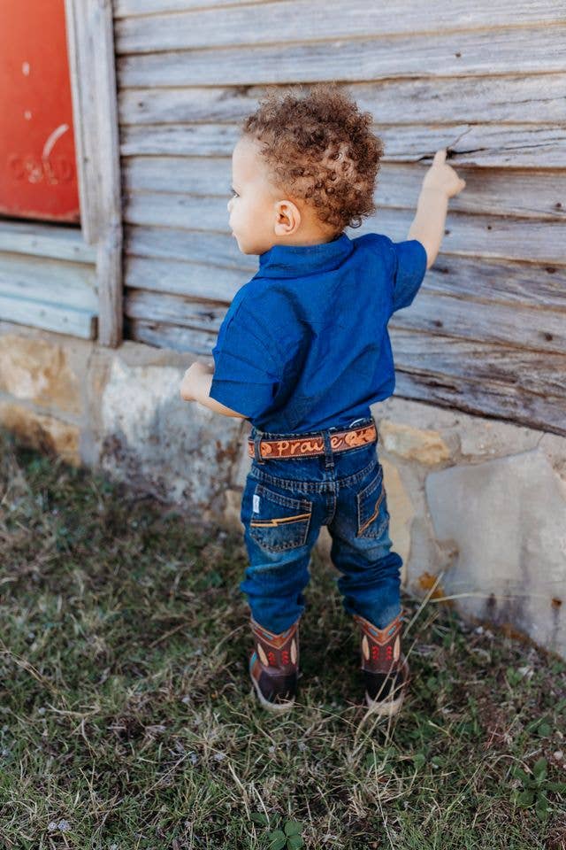 Child in a blue shirt and jeans standing in front of a rustic wooden wall.