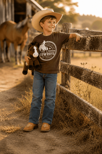 A young boy wearing a cowboy hat, brown graphic t-shirt, blue jeans, and boots stands on a dirt path next to a wooden fence at a sunny outdoor ranch, holding a stuffed animal.