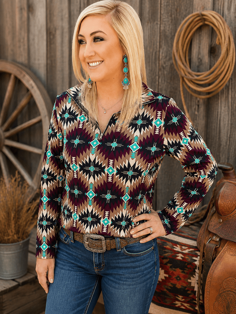 A woman wearing a long-sleeve collared button-up shirt with a Southwestern geometric pattern in black, burgundy, tan, and turquoise, paired with blue jeans and turquoise earrings, standing in front of a rustic wooden wall with a rope coiled on it.