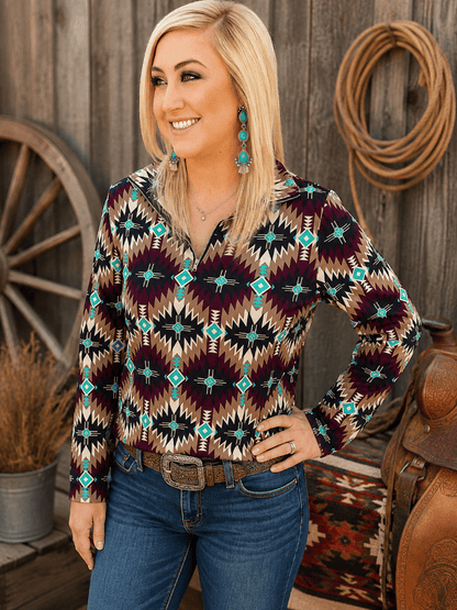 A woman wearing a long-sleeve collared button-up shirt with a Southwestern geometric pattern in black, burgundy, tan, and turquoise, paired with blue jeans and turquoise earrings, standing in front of a rustic wooden wall with a rope coiled on it.