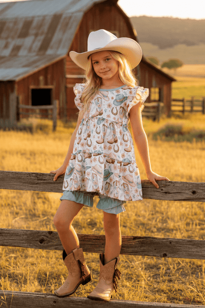 Young girl in a dress with horse print and cowboy boots standing on a wooden fence in a rural setting.