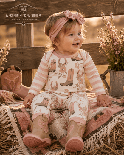 A toddler girl wearing a pink and cream striped long-sleeve romper with cowboy boot prints, matching pants, a pink bow headband, and pink boots, sitting on a patterned blanket in a rustic outdoor setting.