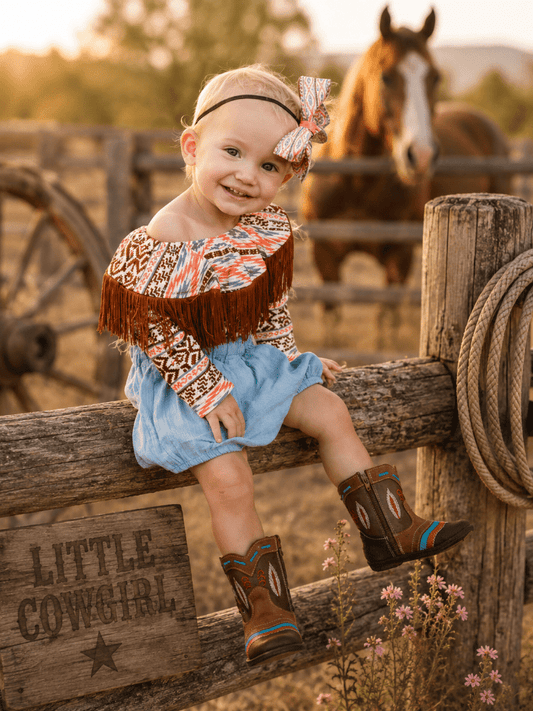 A toddler girl wearing a western-themed cowgirl outfit, including a patterned fringed top, a light blue skirt, and brown cowboy boots, sitting on a wooden fence in a rustic outdoor setting with a horse in the background.