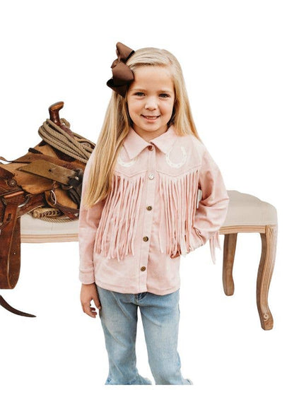 A young girl with long blonde hair wearing a dusty pink western-style fringed shirt and blue jeans, standing in front of a white studio background with a saddle and bench.