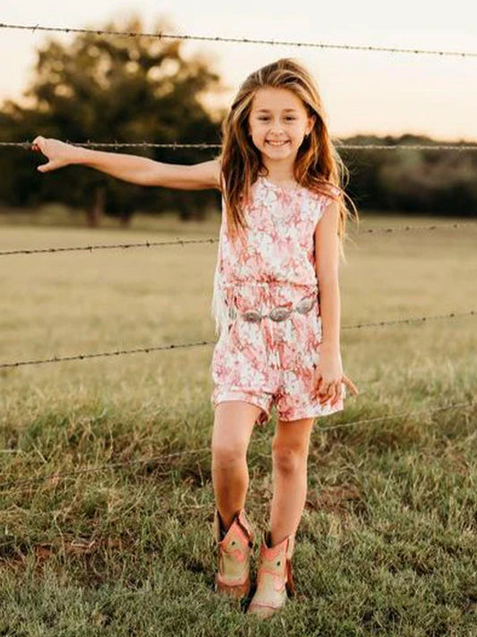 Smiling girl in pink plaid bucking horse fringe romper with cowboy boots, standing by a fence.