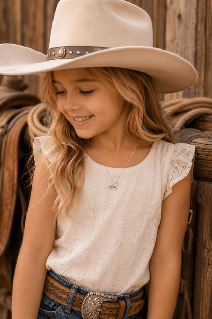 A young girl wearing a cream western-style top with lace ruffle sleeves, a matching cream cowboy hat, and a brown leather belt with a silver concho buckle, posed against a rustic wooden fence background.