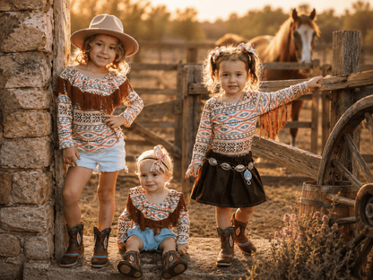 Three young girls in matching western-style fringe tops and boots pose outdoors beside a rustic stone wall and wooden fence, with a horse visible in the background at sunset.