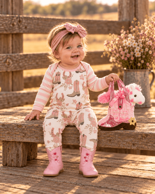 Young baby seated on a rustic bench in a cream and pink cowgirl boot print cotton romper, with full-length legs tucked into Pink Heart baby boots and a pink Giddyup boot purse with plush pony beside her.