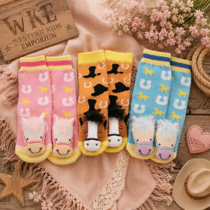 Four pairs of children's novelty socks displayed on a rustic wooden surface with a pink blanket, flowers, rope, and western-themed props. The socks feature horse-themed designs: one pink pair with white horseshoes and plush pink/white horse feet, one orange pair with black horseshoes and brown/white horse faces, one light blue pair with white horseshoes and purple/white horse faces, and one yellow pair with black horseshoes and brown/white horse faces.