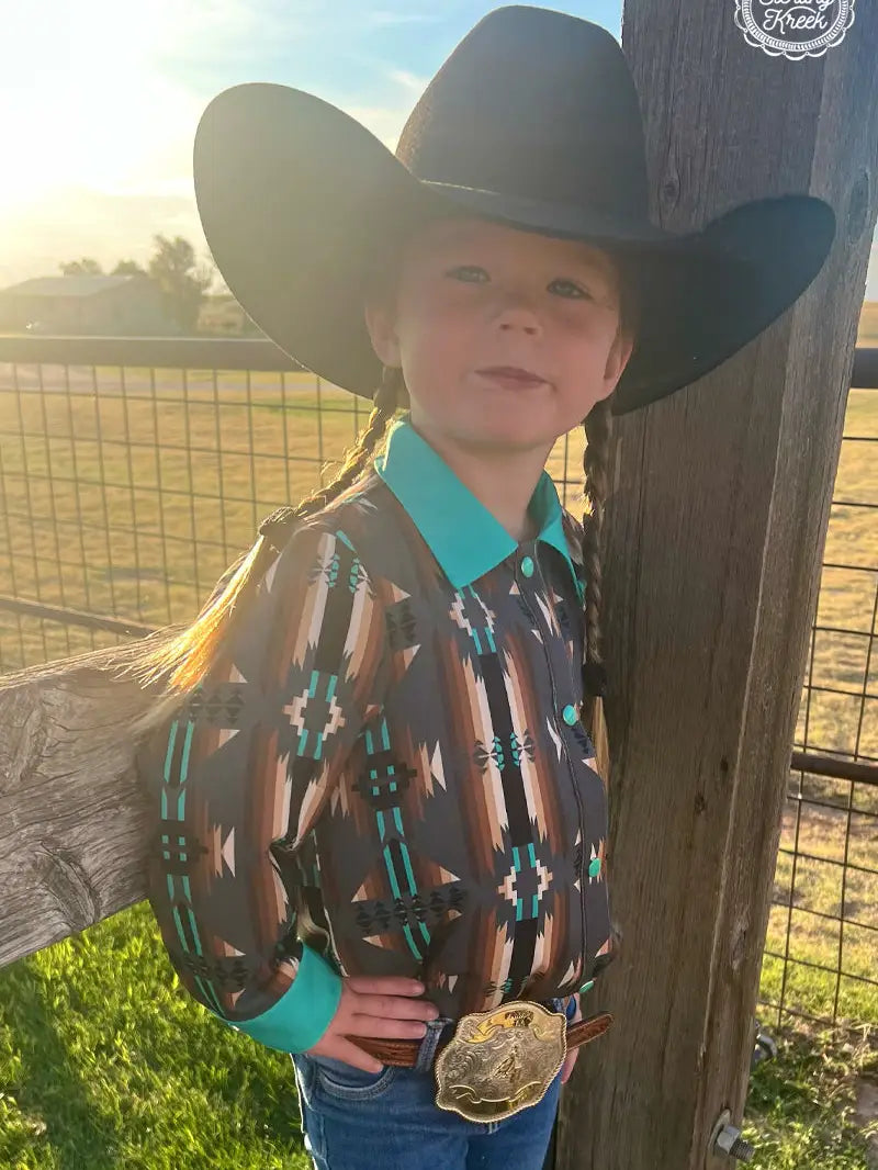 Little girl in an Aztec button-down shirt and turquoise collar leaning on a fence post