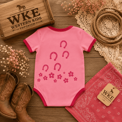 A pink short-sleeve baby bodysuit with dark pink trim and a horseshoe and flower print, styled in a rustic western flat lay with cowboy boots, a WKE Western Kids box, a bandana, rope, and dried flowers on a wooden surface.