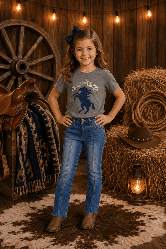 A young girl stands in a rustic barn-like setting wearing a gray graphic T-shirt with a dark blue horse design and the words 'Choose Dream,' paired with blue jeans and brown boots. Hay bales, a wagon wheel, string lights, a saddle, and a lantern are visible in the background.