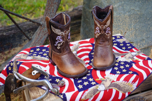 Brown kids' square toe Western boots with cross and wing embroidery on American flag background.