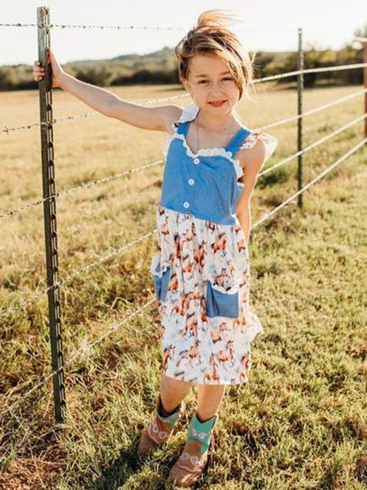 Smiling girl wearing western horse print dress with denim top, standing by a farm fence.