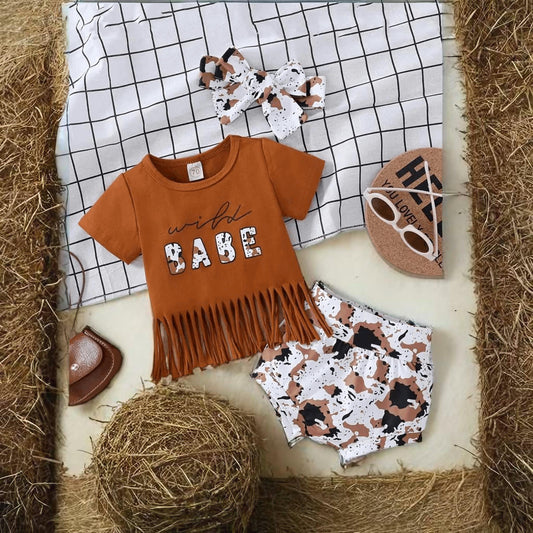 Wild Babe baby outfit flat lay with cow print shorts, tassel top, and matching western accessories for baby girls.